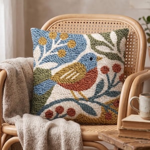 May include: Square decorative pillow with a colorful bird and floral design. The pillow features a blue, green, orange, and white color scheme. The pillow is placed on a wicker chair with a beige blanket draped over it.