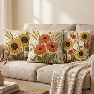 May include: Three square throw pillows featuring textured embroidery of sunflowers, orange poppies, and white daisies on a cream-colored fabric base. The designs showcase detailed floral patterns with green stems and leaves, creating a cohesive botanical set.
