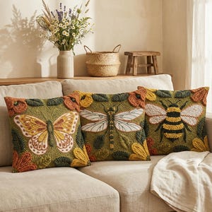 May include: Three decorative throw pillows with embroidered insect designs. One pillow features a butterfly, another a dragonfly, and the third a bee. The pillows have a green background with orange and yellow floral accents, placed on a beige sofa.