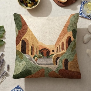 May include: Square decorative pillow with a detailed embroidered design depicting a Mediterranean-style architectural scene in shades of beige, brown, green, and yellow. The pillow is surrounded by olives, bread, and other decorative items.