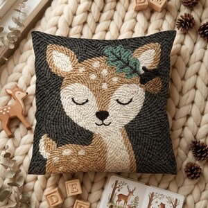 May include: A square throw pillow featuring a punch needle design of a fawn with closed eyes, a white spotted back, and a green leaf on its head, set against a dark charcoal gray background with a textured, looped yarn finish.