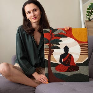 May include: A woman holds a colorful needlepoint artwork depicting a meditating Buddha figure in front of a sunset with a tree. The artwork is framed with a brown border.