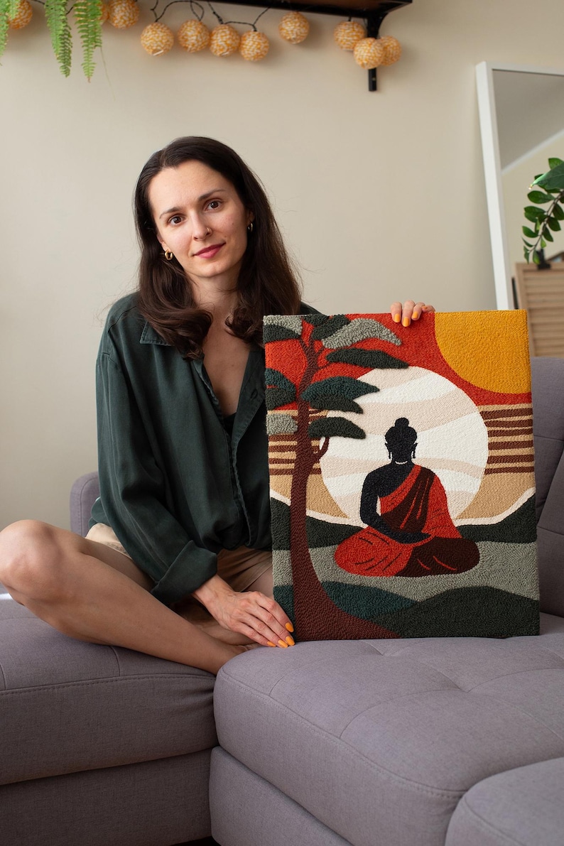 May include: A woman holds a colorful needlepoint artwork depicting a meditating Buddha figure in front of a sunset with a tree. The artwork is framed with a brown border.