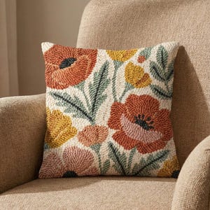 May include: Square cushion with a floral pattern. The cushion displays a cream backdrop with orange, yellow, pink, and green flowers. It is placed on a beige armchair.