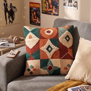 May include: A square throw cushion featuring a textured punch needle design with a geometric pattern. The surface displays a mix of cream, burnt orange, and deep teal yarn in various shapes including diamonds, circles, and triangles.