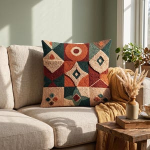 May include: A square throw pillow featuring a textured punch needle design with a geometric pattern. The surface displays a grid of squares in shades of burnt orange, deep teal, and cream, with raised shapes including circles, diamonds, and triangles.