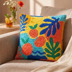 May include: Square decorative pillow with a colorful abstract design. The pillow features a yellow background with blue, orange, and green embroidered shapes resembling leaves and fruit. The pillow is placed on a neutral-toned armchair.