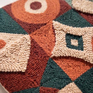 May include: A punch needle textile featuring a geometric pattern of squares and triangles in shades of rust orange, dark forest green, and cream. The design incorporates raised, textured loops of yarn that create a tactile surface across the fabric.