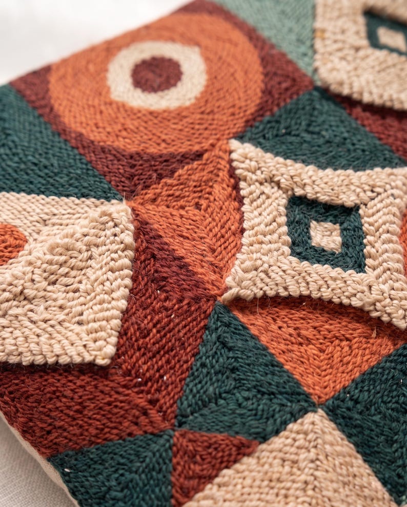 May include: A punch needle textile featuring a geometric pattern of squares and triangles in shades of rust orange, dark forest green, and cream. The design incorporates raised, textured loops of yarn that create a tactile surface across the fabric.