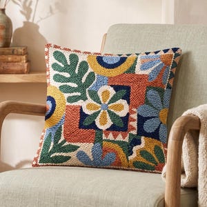May include: A square throw pillow featuring a textured punch needle design with a colorful botanical pattern. The surface displays abstract floral and leaf motifs in shades of mustard yellow, terracotta orange, forest green, and navy blue.