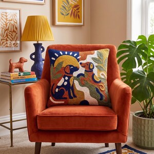 May include: A square throw pillow featuring a textured punch needle design of two dogs in a landscape with a sun, rendered in a palette of navy blue, mustard yellow, terracotta, and cream, resting on a burnt orange velvet armchair.