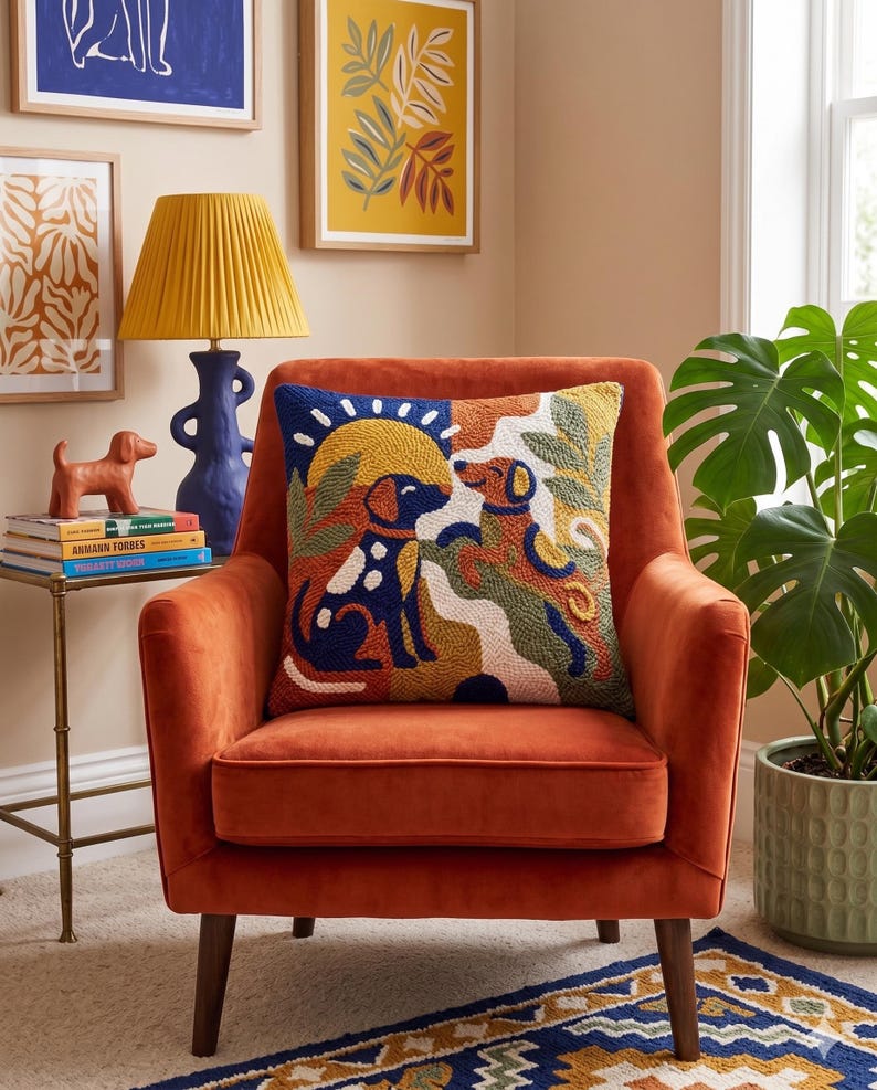 May include: A square throw pillow featuring a textured punch needle design of two dogs in a landscape with a sun, rendered in a palette of navy blue, mustard yellow, terracotta, and cream, resting on a burnt orange velvet armchair.