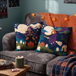 May include: Two square throw pillows featuring punch needle embroidery with Halloween themes. One pillow displays a black cat wearing a purple witch hat holding a pumpkin. The other pillow shows a person flying on a broomstick past a full moon.