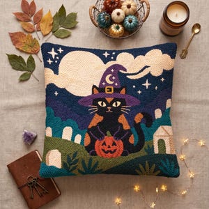 May include: A square throw pillow featuring a punch needle design of a black cat wearing a purple witch hat, holding an orange jack-o'-lantern. The background depicts a night scene with a large cream moon, clouds, stars, and a graveyard.