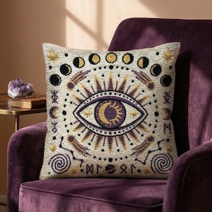 May include: A square throw pillow with a cream-colored base featuring an embroidered design of a central eye with a sun and moon pupil. The pattern includes a lunar phase cycle, golden stars, purple spiral motifs, and celestial symbols.