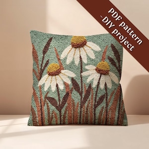 May include: A square, textured pillow with a daisy floral design. The pillow features three large daisies with white petals and yellow centers, set against a green background. Brown and orange grass-like elements surround the flowers. Text on the image reads: PDF pattern - DIY project.