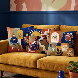 May include: Three square throw pillows featuring textured punch needle embroidery in a vibrant palette of navy blue, mustard yellow, orange, and green. The designs depict stylized scenes of people with pets, dogs playing under a sun, and paw prints.