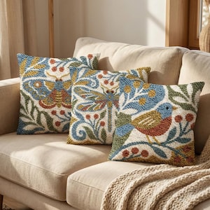 Op de afbeelding: Drie sierkussens met geborduurde ontwerpen van insecten en vogels. De kussens liggen op een beige bank met een bijpassende deken. De ontwerpen bevatten blauwe, groene, rode en gele accenten.