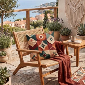 May include: A wooden armchair with a woven cane back and seat, featuring a square throw pillow with a geometric punch needle pattern in shades of rust, teal, and cream, draped with a textured rust-coloured throw blanket on the seat.