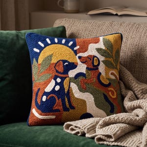 May include: A square throw pillow featuring a textured punch needle design of two dogs in a landscape with a yellow sun. The color palette includes deep blue, burnt orange, mustard yellow, olive green, and cream, with a dark blue piped edge.