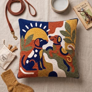 May include: A square punch needle throw pillow featuring a colorful graphic of two dogs, one blue and one brown, set against a yellow sun and green foliage. The design uses textured yarn loops in shades of blue, yellow, orange, green, and white.
