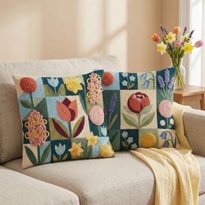 May include: Two decorative throw pillows with embroidered floral designs. The pillows feature a patchwork pattern with various flowers in shades of orange, yellow, pink, and blue on a dark teal background. The pillows are placed on a beige sofa.