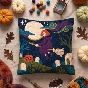 May include: A square punch needle pillow featuring a person with red hair flying on a broomstick against a large yellow moon. The scene includes a dark blue night sky, purple clouds, a jack-o'-lantern, gravestones, and a small house.