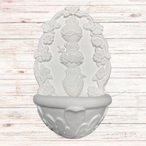 May include: White wall-mounted fountain with ornate floral and vase detailing. The fountain features a basin at the bottom and a decorative arched top with flowers. The central design includes a vase with floral accents and a cross.