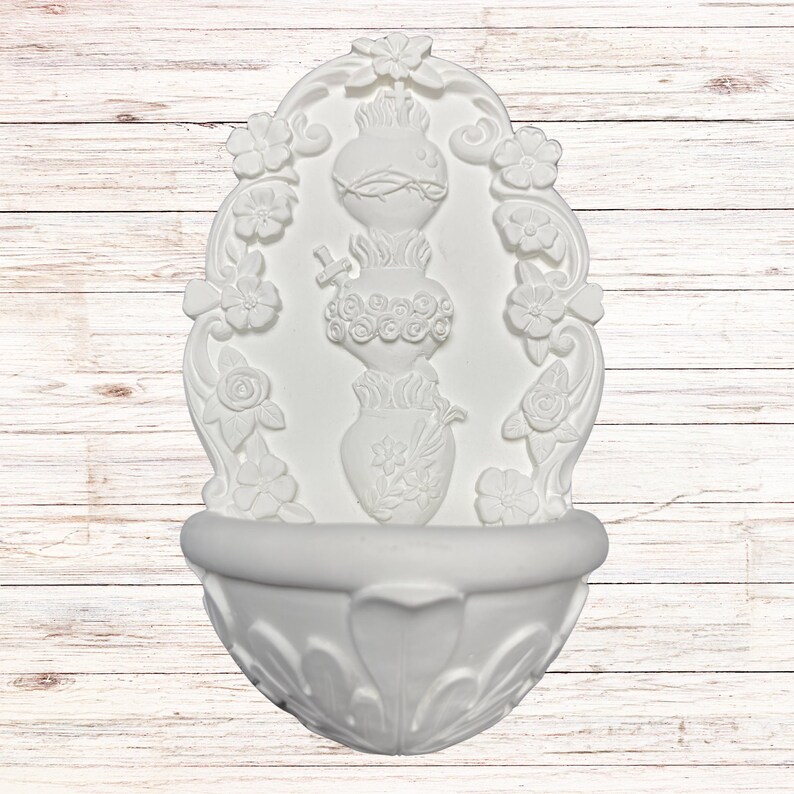 May include: White wall-mounted fountain with ornate floral and vase detailing. The fountain features a basin at the bottom and a decorative arched top with flowers. The central design includes a vase with floral accents and a cross.