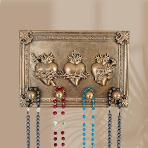 May include: A rectangular bronze-toned wall plaque featuring three raised heart motifs, each topped with flames. The left heart is pierced by a sword, the center is encircled by thorns, and the right is adorned with flowers. Four brass hooks hold beaded necklaces.