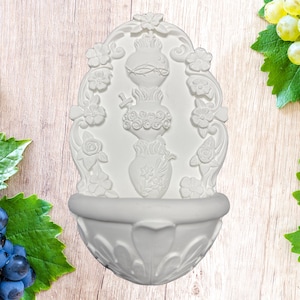 May include: A white, ornate wall fountain with floral and heart-shaped details. The fountain features a basin at the bottom and a decorative top with a central design of a heart and flowers. The overall design is symmetrical and detailed.