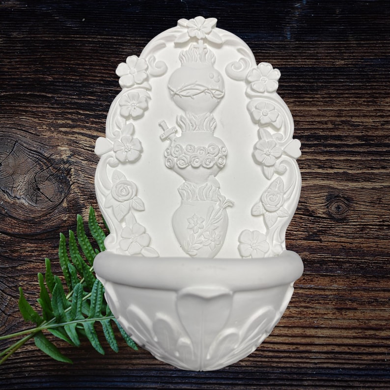 May include: White wall-mounted fountain with floral and heart-shaped relief details. The fountain features a basin at the bottom and a decorative top with three stacked vase-like elements. The design includes flowers and a fern leaf.