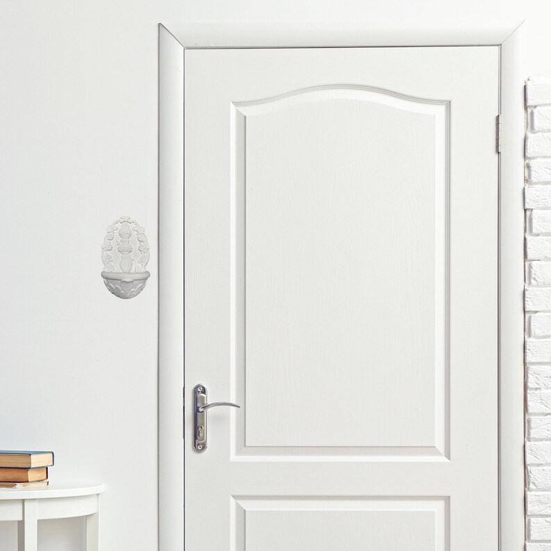 May include: A white wall-mounted decorative sculpture featuring a floral design. The sculpture is mounted on a white wall next to a white door. The door has a silver handle and a simple design.