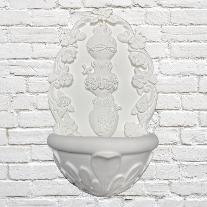 May include: White wall-mounted decorative fountain with floral and vase motifs. The fountain features an oval frame with flowers, a central vase design, and a lower basin with a heart-shaped detail. The background is a white brick wall.