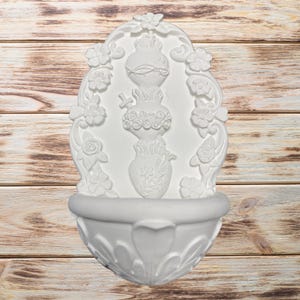 May include: White wall-mounted decorative fountain with floral and ornate details. The fountain features a basin at the bottom and a central design with a vase and floral accents. The background is a light brown wooden surface.