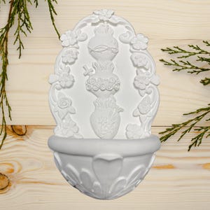 May include: White wall-mounted decorative fountain with floral and heart-shaped relief details. The fountain features a basin at the bottom and a central design with a heart, flowers, and a cross. The design is surrounded by floral embellishments.