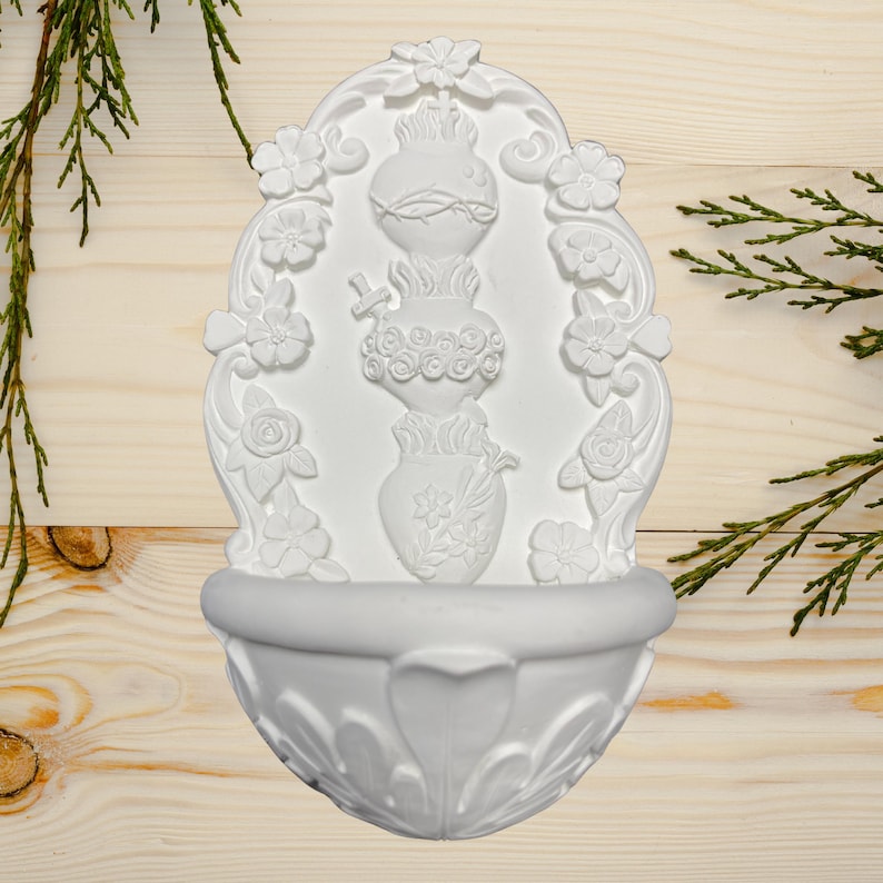 May include: White wall-mounted decorative fountain with floral and heart-shaped relief details. The fountain features a basin at the bottom and a central design with a heart, flowers, and a cross. The design is surrounded by floral embellishments.