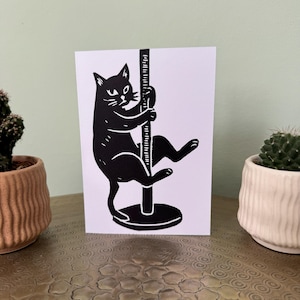 May include: A white greeting card featuring a black ink linocut illustration of a cat climbing a vertical pole. The cat is depicted in profile with its front paws gripping the pole and its hind legs extended, set against a plain white background.