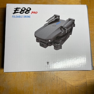 May include: A gray foldable drone on a white box. The box has the text "E88 PRO FOLDABLE DRONE" in black and red. The drone is dark gray with black propellers and a camera.