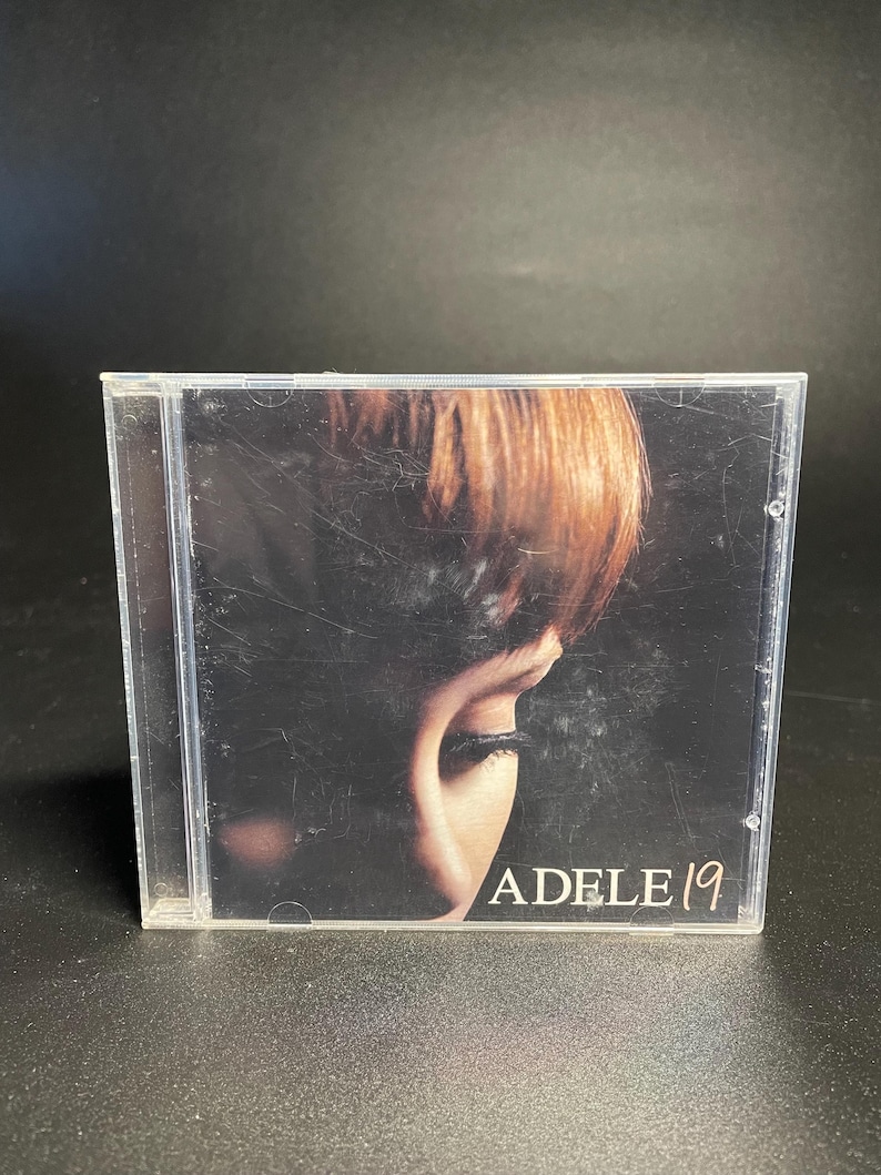 May include: A CD case with a close-up image of a woman's face. The text "ADELE 19" is printed on the front of the case.