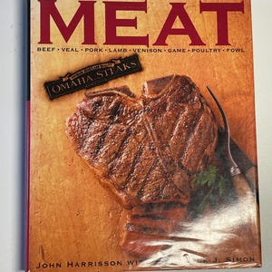 Może przedstawiać: Książka kucharska zatytułowana "MEAT" z zbliżeniem grillowanego steku na okładce. Podtytuł brzmi "BEEF VEAL PORK LAMB VENISON GAME POULTRY FOWL". Na okładce widoczna jest czerwono-biała etykieta z tekstem "PREMIUM HEARTLAND QUALITY OMAHA STEAKS SINCE 1917".