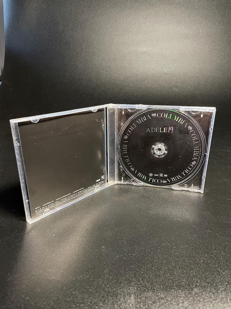 May include: A clear plastic CD case with a black CD inside. The CD has the text "Adele 19" and "Columbia" printed on it.