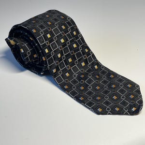 May include: A rolled-up black tie with a repeating geometric pattern of squares, circles, and rectangles in white, yellow, and blue. The tie is made of a smooth, possibly silk, material.