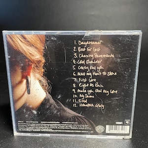 May include: The back of a CD case with a black and white photo of a woman with a large earring. The CD case lists the track titles for the album "19" by Adele. The track titles are: Daydreamer, Best For Last, Chasing Pavements, Cold Shoulder, Crazy for You, Melt My Heart To Stone, First Love, Right As Rain, Make You Feel My Love, My Same, Tired, Hometown Glory.