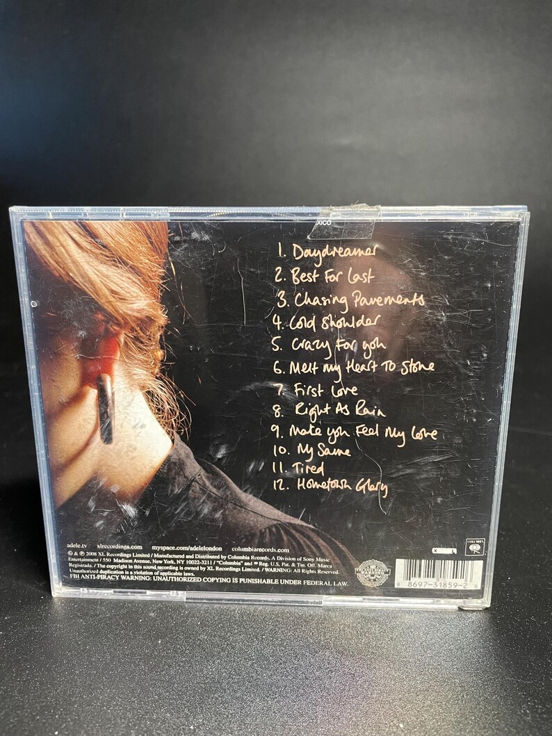 May include: The back of a CD case with a black and white photo of a woman with a large earring. The CD case lists the track titles for the album "19" by Adele. The track titles are: Daydreamer, Best For Last, Chasing Pavements, Cold Shoulder, Crazy for You, Melt My Heart To Stone, First Love, Right As Rain, Make You Feel My Love, My Same, Tired, Hometown Glory.