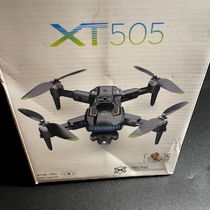 Op de afbeelding: Een zwarte en grijze XT 505 drone met vier propellers en een camera wordt getoond op een witte doos. De doos heeft de modelnaam van de drone in blauw en groen. De drone heeft groene accenten op de propellers.