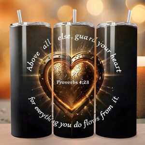 以下が含まれることがあります： 黒い背景に金色のハートが描かれたステンレス製のタンブラー。"Above all else, guard your heart for everything you do flows from it."と"Proverbs 4:23"の文字が印刷されています。