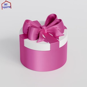 Puede incluir: Una caja de regalo redonda con tapa blanca y base rosa brillante. Un gran lazo rosa a juego se encuentra encima de la tapa. La caja se presenta sobre un fondo blanco.