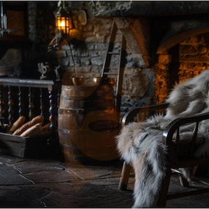 May include: A rustic wooden chair with a faux fur throw draped over it sits in front of a stone fireplace with a roaring fire. The chair is in a dimly lit room with wooden floors and a barrel in the background.