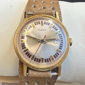 Könnte beinhalten: Vintage Timex-Uhr mit goldfarbenem Gehäuse und silbernem Zifferblatt. Die Uhr hat ein braunes Lederarmband mit Perforationen. Das Zifferblatt zeigt den Markennamen und eine Datumsanzeige. Die Uhr wird auf einer weißen Oberfläche präsentiert.